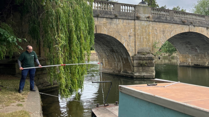 A photo of a man in a green top who is holding a long pole over the River Thames at Wallingford Bridge. String from the pole goes down into the water where a testing tube is collecting water samples.